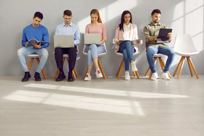 Smart Young People Using Laptops, Digital Tablets And Textbooks Sitting In Row In Waiting Room.